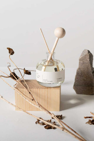 Hetkinen Co Kalevala reed diffuser from Finland in a minimalist glass bottle with light wood reeds, placed on a wooden block beside dried branches, leaves, and a Finnish granite stone on a white background.