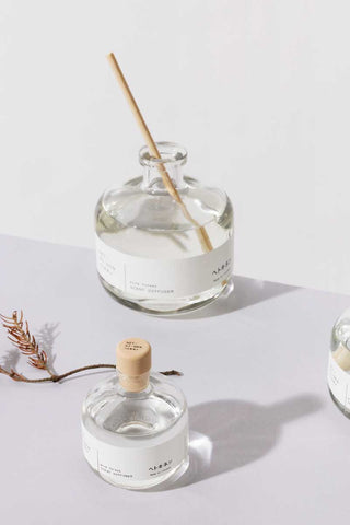 Hetkinen Co Pine Forest reed diffuser from Finland in minimalist glass bottles with light wood reeds, shown in an open and closed bottle arrangement with a dried plant accent, styled with soft shadow play.