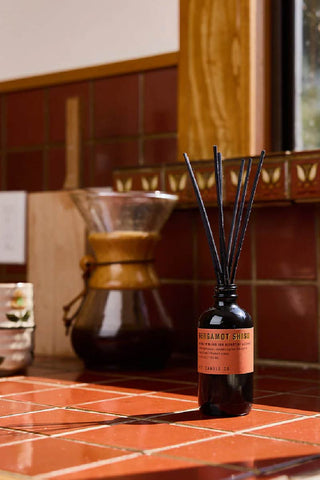 P.F. Candle Co Bergamot Shiso reed diffuser with reed sticks in a 103ml glass bottle on a maroon cottagecore tabletop, placed beside a sunflower-painted cup