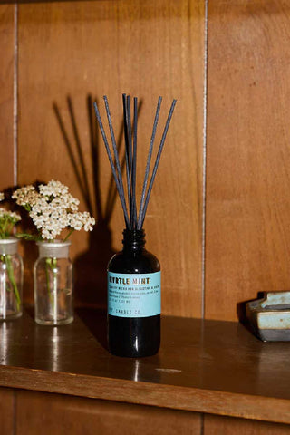 p.f. candle co myrtle mint reed diffuser with reed sticks in 103ml glass bottle on a retro wood shelf next to two glass vase of small white flowers fresh and earthy home fragrance

