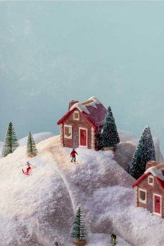 Paddywax Alpine Chalet incense houses styled in a snowy mountain village scene with pine trees and miniature skiers, red-roof log cabins evoking cozy winter chalet vibes, festive home fragrance décor for gifting.