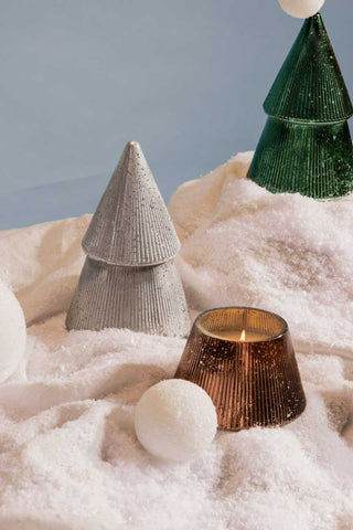 Paddywax Cypress & Fir silver and green metallic tree candles displayed on snowy fabric with glowing bronze candle, cozy festive Christmas décor and holiday candle gift inspiration.