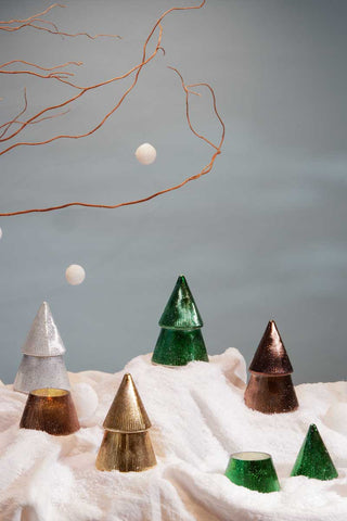 Paddywax Cypress & Fir tree candles in green, gold, silver, and bronze metallic glass arranged on snowy fabric with branch and floating snowballs, festive holiday home décor and Christmas candle gift inspiration.