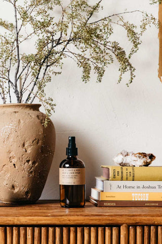 P.F. Candle Co Amber and Moss room spray placed on a fluted wooden tabletop beside a stack of books topped with a rock, styled to evoke an earthy and fresh home atmosphere.
